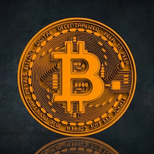 Op de afbeelding: Een goudkleurige Bitcoin-munt met het Bitcoin-symbool in het midden en de tekst "DIGITAL DECENTRALIZED PEER TO PEER" rond de rand. De munt heeft ook de tekst "MJB MONETARY METALS BITCOIN" en "TAYO 999 FINE COPPER" op de rand.