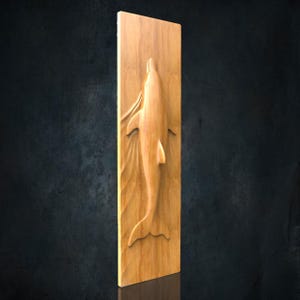 May include: A rectangular light brown wooden wall plaque featuring a carved relief of a dolphin swimming vertically. The dolphin is depicted in a smooth, polished finish, with carved wave patterns surrounding its body on the flat wooden surface.