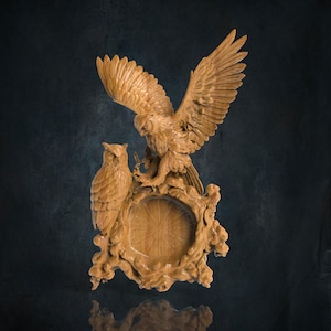 Pode incluir: Uma escultura de madeira de uma águia e uma coruja empoleiradas em uma moldura circular com detalhes intrincados. A águia tem as asas abertas, enquanto a coruja olha para o lado. A moldura é de madeira e tem um acabamento natural.