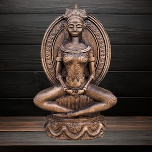 May include: A detailed wooden statue of a seated figure in a meditative pose. The figure has intricate carvings, wears a crown, and is surrounded by an oval halo. The statue is a warm brown color and sits on a decorative base.