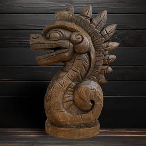 May include: A detailed wooden sculpture of a mythical creature with an open mouth and a feathered headdress. The sculpture is brown and stands on a round base. The background is a dark wooden wall.