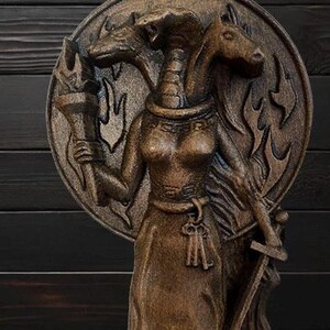 May include: A wooden sculpture of a three-headed goddess with a torch in one hand and a sword in the other. The goddess is wearing a long robe and a crown. The sculpture is painted in shades of brown and black.