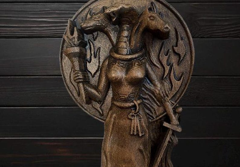 May include: A wooden sculpture of a three-headed goddess with a torch in one hand and a sword in the other. The goddess is wearing a long robe and a crown. The sculpture is painted in shades of brown and black.