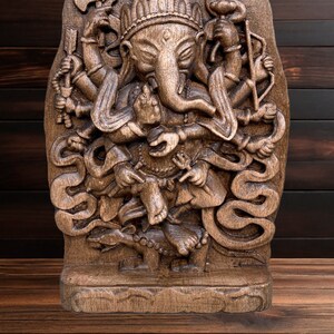 May include: A detailed wooden carving of the Hindu deity Ganesha, with multiple arms and a trunk, standing on a snake. The carving is dark brown and has a textured surface.