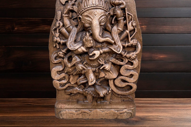 May include: A detailed wooden carving of the Hindu deity Ganesha, with multiple arms and a trunk, standing on a snake. The carving is dark brown and has a textured surface.