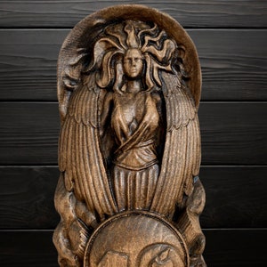 May include: A wooden carving of a winged figure with flowing hair and a shield. The figure is carved in a detailed, realistic style and has a warm brown colour.