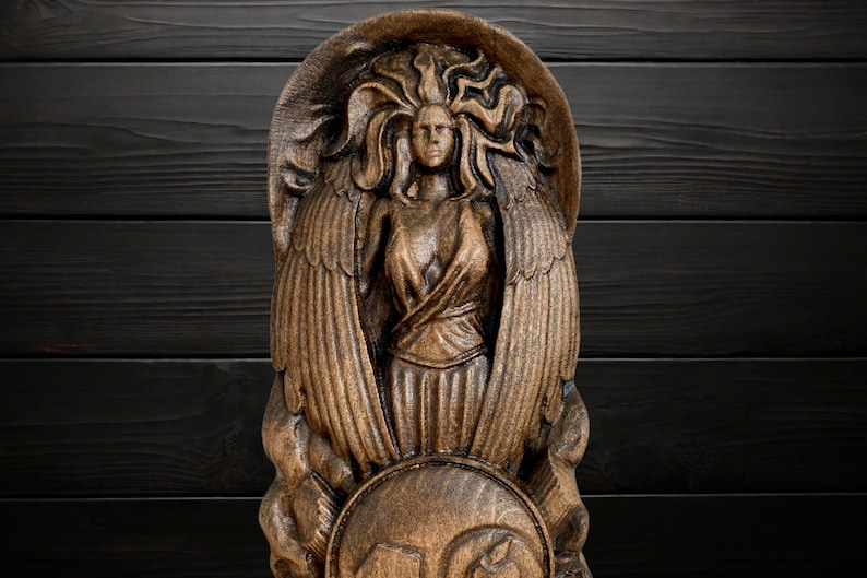 May include: A wooden carving of a winged figure with flowing hair and a shield. The figure is carved in a detailed, realistic style and has a warm brown colour.
