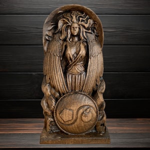 May include: A wooden sculpture of a winged female figure holding a shield with a stylized design. The sculpture is carved in a dark brown wood and has a detailed, intricate design.