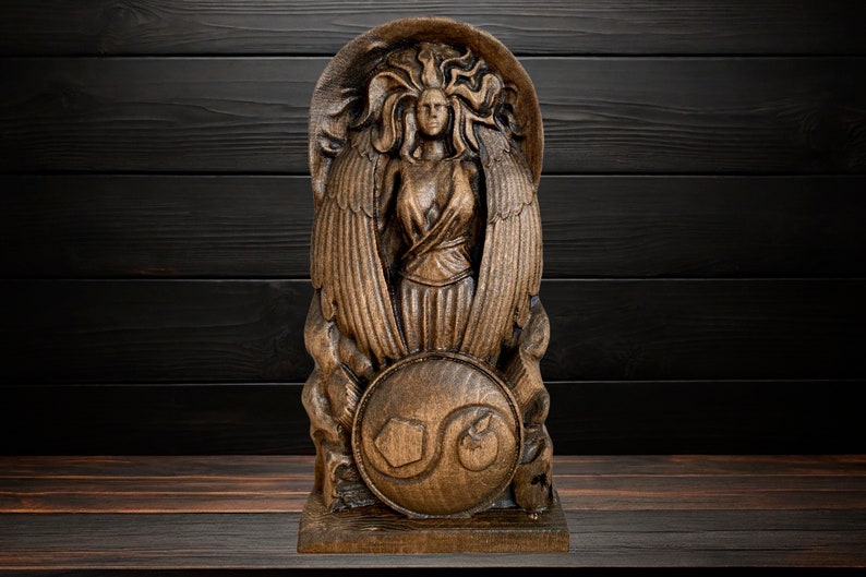 May include: A wooden sculpture of a winged female figure holding a shield with a stylised design. The sculpture is carved in a dark brown wood and has a detailed, intricate design.