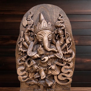 May include: A detailed wooden carving of the Hindu deity Ganesha, with multiple arms and a trunk, standing on a snake. The carving is set against a dark wood background.