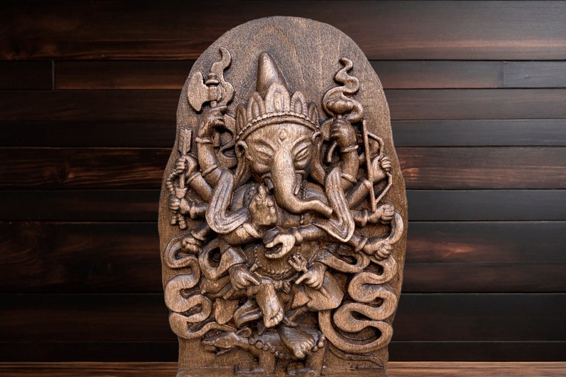 May include: A detailed wooden carving of the Hindu deity Ganesha, with multiple arms and a trunk, standing on a snake. The carving is set against a dark wood background.