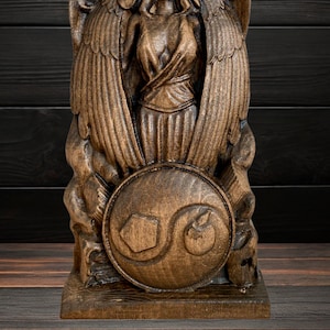 May include: A wooden sculpture of a winged figure holding a shield with a carved design. The sculpture is brown and has a detailed finish.