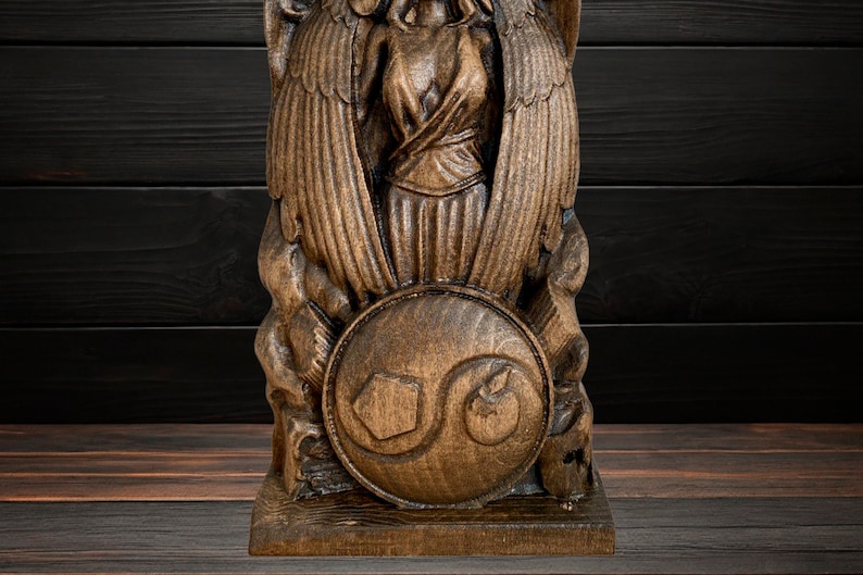 May include: A wooden sculpture of a winged figure holding a shield with a carved design. The sculpture is brown and has a detailed finish.