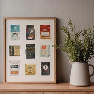 May include: A framed print featuring watercolor illustrations of various coffee bags. The bags are in different colors and designs, with text labels. The print is displayed on a wooden surface, with a white pitcher holding greenery to the right.