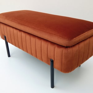 Op de afbeelding: Een bruine fluwelen opbergbank met zwarte metalen poten. De bank heeft een gestoffeerde bovenkant en een rechthoekige vorm.