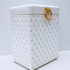 Peut inclure: Boîte de rangement matelassée blanche avec une poignée en anneau dorée et des clous dorés.