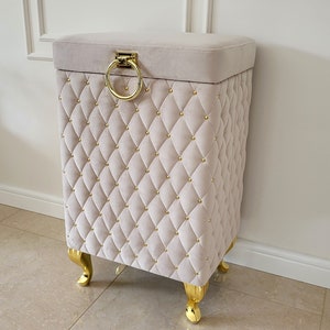 Peut inclure: Un pouf de rangement de couleur crème avec un design capitonné et des pieds dorés. Le pouf a une poignée en forme d'anneau dorée sur le dessus.