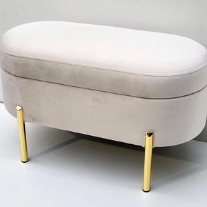Könnte beinhalten: Ein beige gepolsterter Hocker mit abgerundeter rechteckiger Form. Der Hocker hat eine weiche, samtige Textur und wird von vier goldfarbenen Metallbeinen getragen. Die Oberseite ist ein hellerer Beigeton als die Basis.