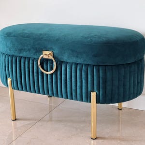 Puede incluir: Otomana de terciopelo color turquesa con patas de metal dorado y un tirador de anillo dorado a juego. El banco de almacenamiento ovalado presenta detalles acanalados verticales en la base y una parte superior lisa y acolchada. Un mueble elegante y funcional.