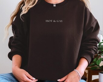 May include: A dark brown sweatshirt with the text "HOT & GAY" in white. The sweatshirt is a crew neck style and is being worn by a person wearing blue jeans and white sneakers.