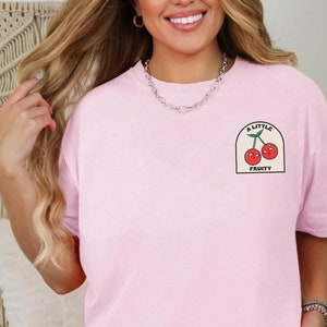 May include: A pink t-shirt with a graphic of two red cherries with faces and the text "A Little Fruity" on a white patch.