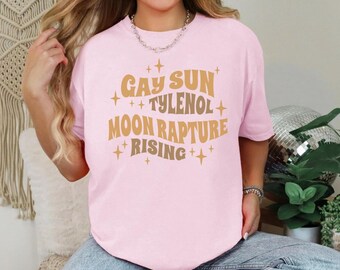 May include: A light pink t-shirt with the words "GAY SUN TYLENOL MOON RAPTURE RISING" in a gold and brown font. The text is surrounded by small star shapes. The shirt is a casual style.