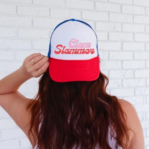 May include: A white, red, and blue trucker hat with a mesh back. The hat has the text "Clam Slammer" in pink and red letters.