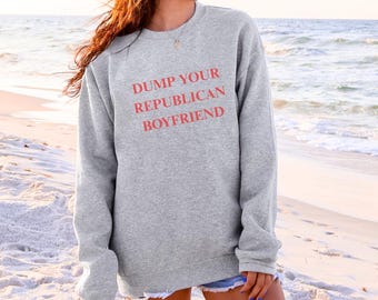 May include: A gray sweatshirt with the text "DUMP YOUR REPUBLICAN BOYFRIEND" in red. The sweatshirt is a crew neck style and is worn on a beach.