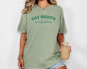 May include: Sage green t-shirt with the text "GAY RIGHTS" in green, with the phrase "as in I'm gay and I'm right" underneath. The shirt is paired with denim shorts. A glass with a straw and a beige drink is held in the hand.