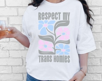 May include: White t-shirt with the text "Respect My Trans Homies" in gray. The shirt features a floral design in light blue and pink, with gray leaves. The shirt has short sleeves.