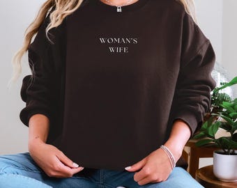 May include: A dark brown sweatshirt with the text "WOMAN'S WIFE" in white. The sweatshirt is a crew neck style and is made of a soft material. The model is wearing blue jeans and white sneakers.