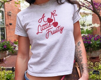 May include: A light gray crop top with the text "A Little Fruity" in red script, featuring a cherry graphic. The shirt is short-sleeved and has a crew neck. The model is wearing blue jeans.