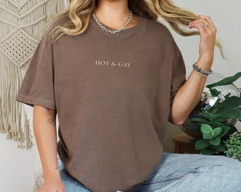May include: A brown, short-sleeved t-shirt with the text "HOT & GAY" printed in white. The shirt is made of a soft material and has a relaxed fit. The shirt is a unisex style.