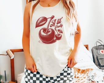 May include: Cream-colored tank top with a red cherry graphic and the text "A lil' Fruity." The top is paired with black and white checkered shorts. The image captures a casual, summery aesthetic.