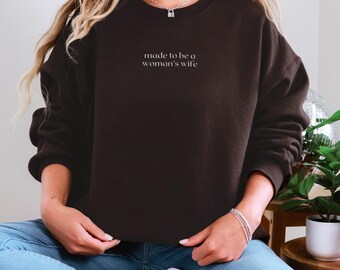 May include: A dark brown sweatshirt with the text "made to be a woman's wife" in a white, minimalist font. The sweatshirt is worn with blue jeans and white sneakers. A silver necklace and bracelet are also visible.