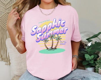 May include: A light pink t-shirt with the words "Sapphic Summer" in a retro font, featuring palm trees and the text "Sun, Salt, Sand, and Shimmer." The shirt has a relaxed fit.