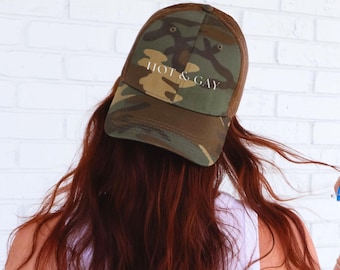 May include: A camouflage baseball cap with the text "HOT & GAY" in white lettering. The cap features a brown mesh back and a curved brim. The front of the cap has a green, brown, and tan camouflage pattern.