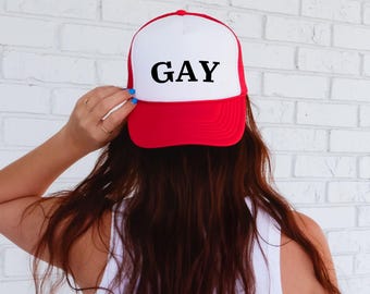 May include: A red and white trucker hat with the word "GAY" printed in black across the front. The hat has a mesh back and a curved brim. The hat is being held by a person.