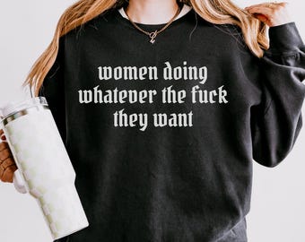 May include: Black sweatshirt with the text "women doing whatever the fuck they want" in white gothic font. The sweatshirt has long sleeves and a crew neck. A white and green checkered tumbler is held in the left hand.