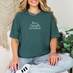 May include: A dark teal t-shirt with the words "live laugh scissor" in white text. The shirt has short sleeves and a relaxed fit. The model is wearing blue jeans and white sneakers.