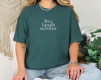 May include: A dark teal t-shirt with the words "live laugh scissor" in white text. The shirt has short sleeves and a relaxed fit. The model is wearing blue jeans and white sneakers.