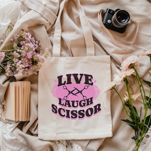 May include: A cream-colored tote bag with the words "LIVE LAUGH SCISSOR" in black and pink. The design includes crossed scissors. The bag is surrounded by flowers, a camera, and a stack of cards.