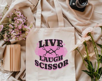 May include: A cream-colored tote bag with the words "LIVE LAUGH SCISSOR" in black and pink. The design includes crossed scissors. The bag is surrounded by flowers, a camera, and a stack of cards.