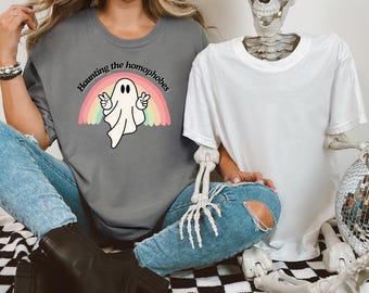 May include: A dark gray t-shirt featuring a white ghost graphic making peace signs in front of a pastel rainbow. The text "Haunting the homophobes" is printed in a black arched font above the ghost. The shirt is paired with blue jeans.