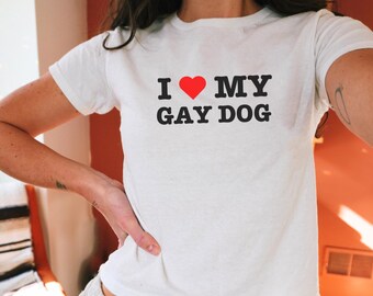 May include: White, short-sleeved t-shirt with the text "I ❤️ MY GAY DOG" in black letters and a red heart. The shirt is a cropped style.