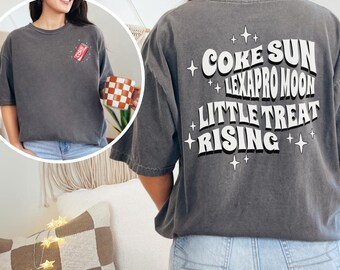 May include: Dark gray t-shirt with the words "COKE SUN LEXAPRO MOON LITTLE TREAT RISING" printed on the back in white. A small "COKE" logo is on the front. The shirt is worn by a person.