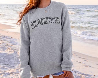 May include: A heather gray sweatshirt with the word "SPORTS" in black block letters across the chest. The sweatshirt has a crew neck and long sleeves. The background is a beach scene with sand and ocean.