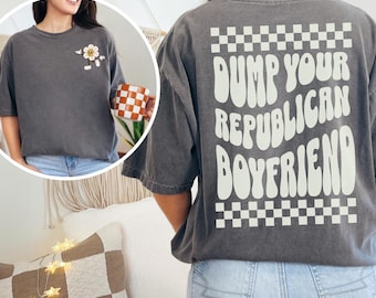 May include: Dark gray t-shirt with the text "DUMP YOUR REPUBLICAN BOYFRIEND" in a retro font on the back. The shirt has a checkerboard pattern at the top and bottom of the text. A smiling flower graphic is on the front.
