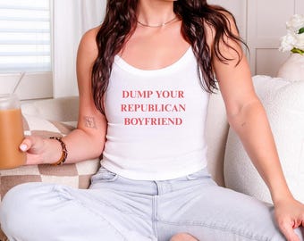 May include: White tank top with the text "DUMP YOUR REPUBLICAN BOYFRIEND" in red. The top is a ribbed style with spaghetti straps. The person is wearing light blue jeans and sitting on a beige couch.