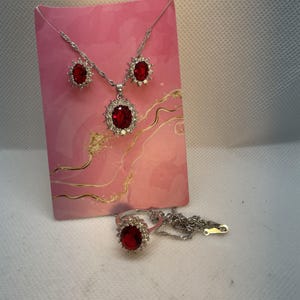 May include: A silver-toned jewelry set featuring a necklace, earrings, and a ring. Each piece has a large, oval, deep red gemstone surrounded by smaller, clear stones. The set is displayed on a pink card.